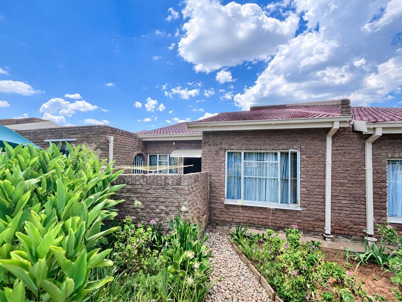 Rear view of a face brick townhouse featuring a tiled roof, aluminium window frames with security bars, and a landscaped garden with a paved pathway.