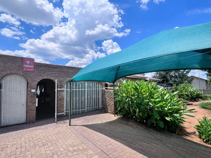 Front exterior view of the townhouse featuring a paved driveway, shade sail carport, and arched entrance with security gate.