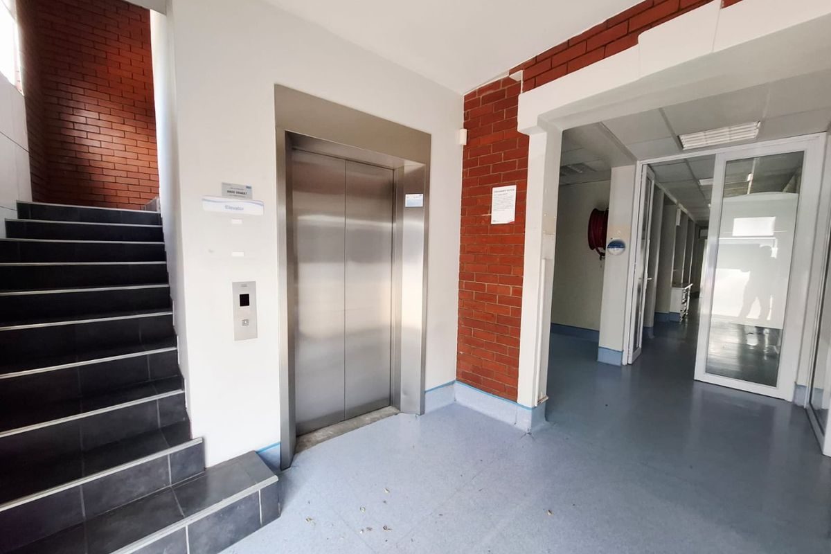 Commercial hallway featuring a stainless steel elevator, staircase with tiled steps, exposed brick accents, and access to adjacent office spaces via glass doors.