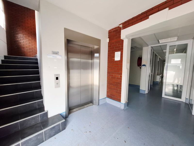 Commercial hallway featuring a stainless steel elevator, staircase with tiled steps, exposed brick accents, and access to adjacent office spaces via glass doors.