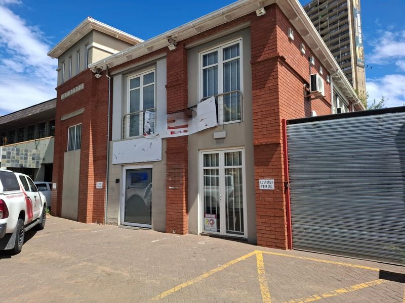 Front exterior view of a commercial brick building featuring a roller shutter door, paved parking area, and balcony access.