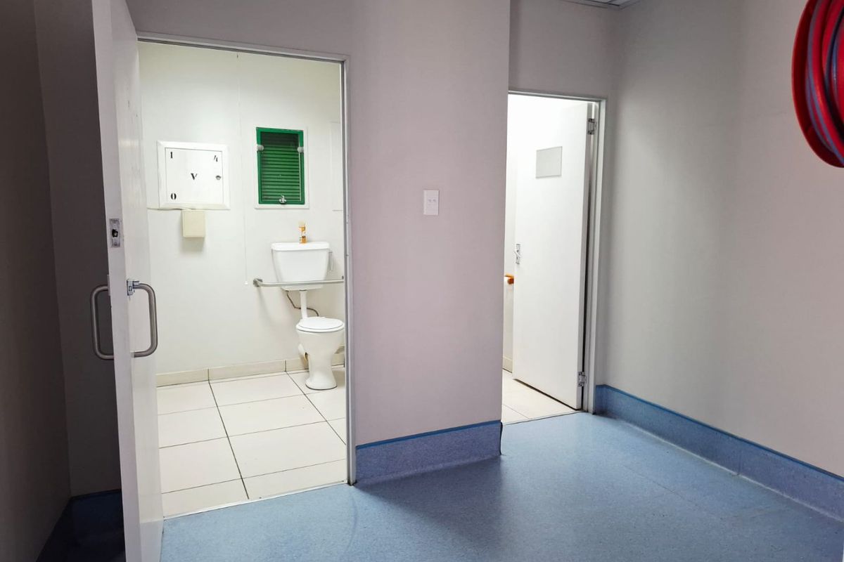 Commercial bathroom facility featuring tiled flooring, a standard toilet, and adjacent utility space with vinyl flooring and fire safety equipment.