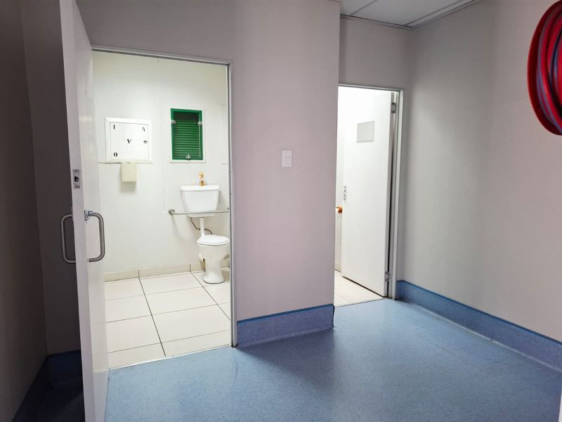 Commercial bathroom facility featuring tiled flooring, a standard toilet, and adjacent utility space with vinyl flooring and fire safety equipment.