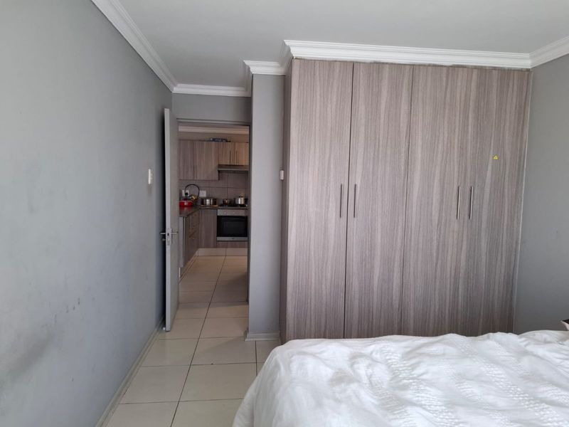 Bedroom featuring built-in cupboards, tiled flooring, and neutral grey walls, with an adjacent view into the kitchen area.