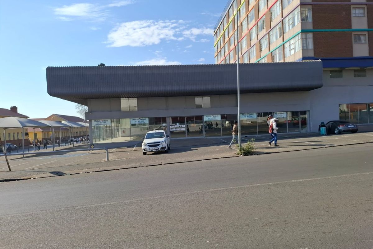 Street-level view of a commercial property featuring a large glass storefront, metal fascia signage, and an adjacent paved parking area.