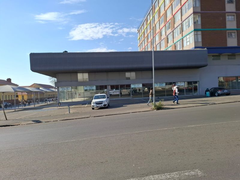 Street-level view of a commercial property featuring a large glass storefront, metal fascia signage, and an adjacent paved parking area.