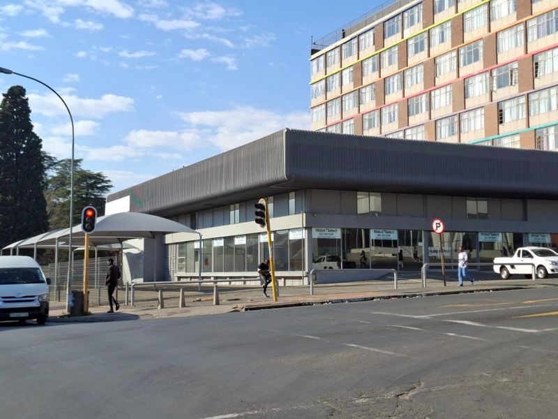 Street-level view of a commercial building featuring large glass storefront windows, metal cladding, and an adjacent shade sail structure.