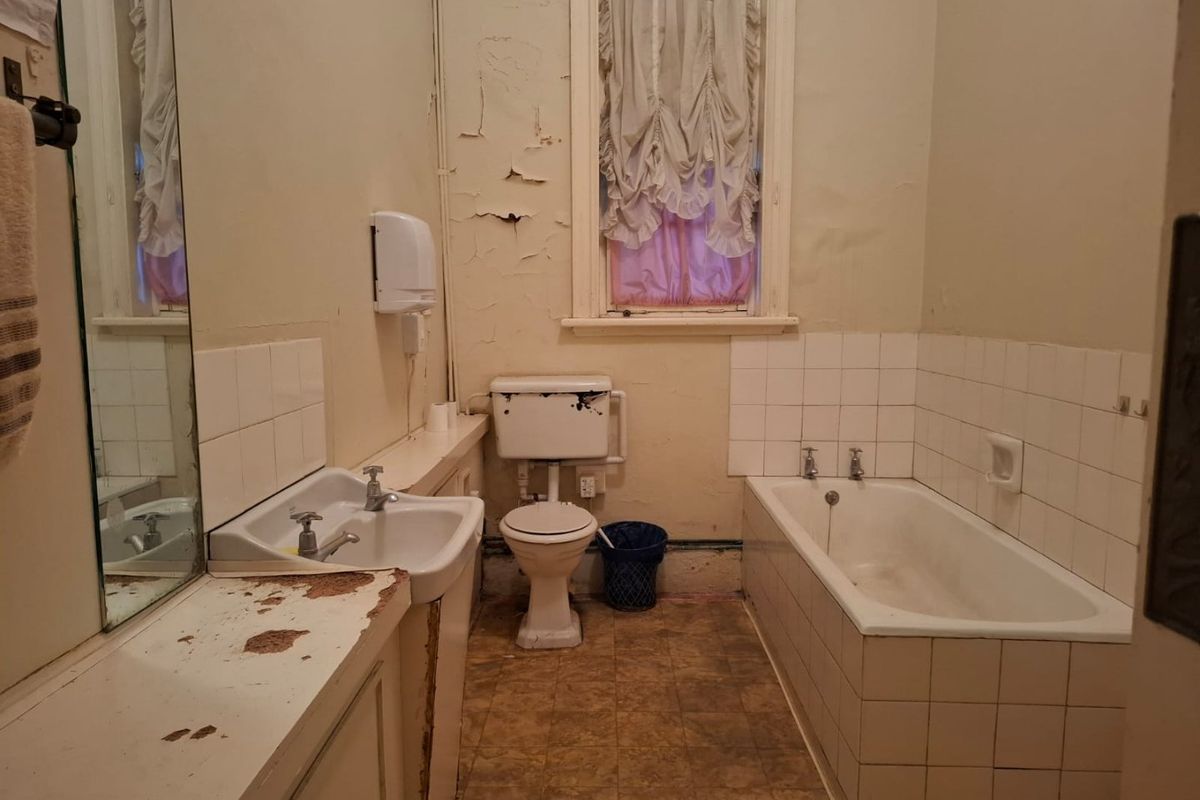 A dated bathroom featuring a built-in vanity, bathtub, and toilet, currently requiring significant cosmetic renovation and repair to wall finishes and surfaces.