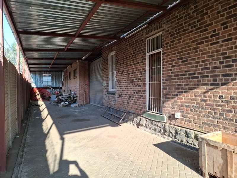 A rear exterior area featuring a covered walkway, brick facade, roller shutter door, and paved flooring, currently used for storage.