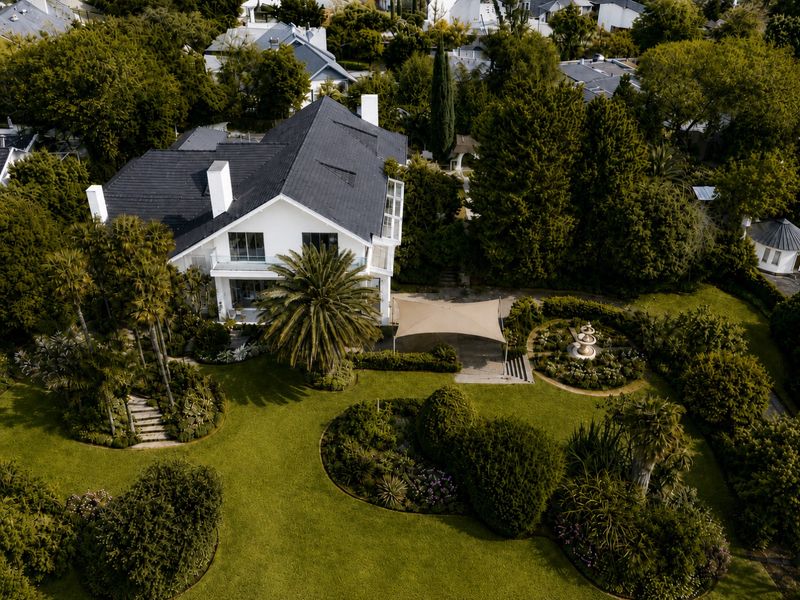 Aerial view of a well-maintained residential property featuring a large landscaped garden, stone fountain, shade sail, and a multi-level house with a tiled roof.