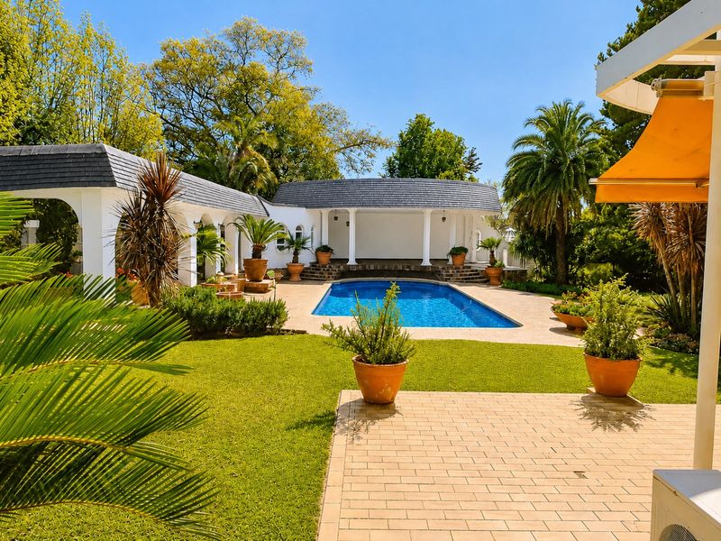 A beautifully landscaped outdoor pool area featuring a sparkling swimming pool, paved patio, and elegant covered walkways with architectural columns.