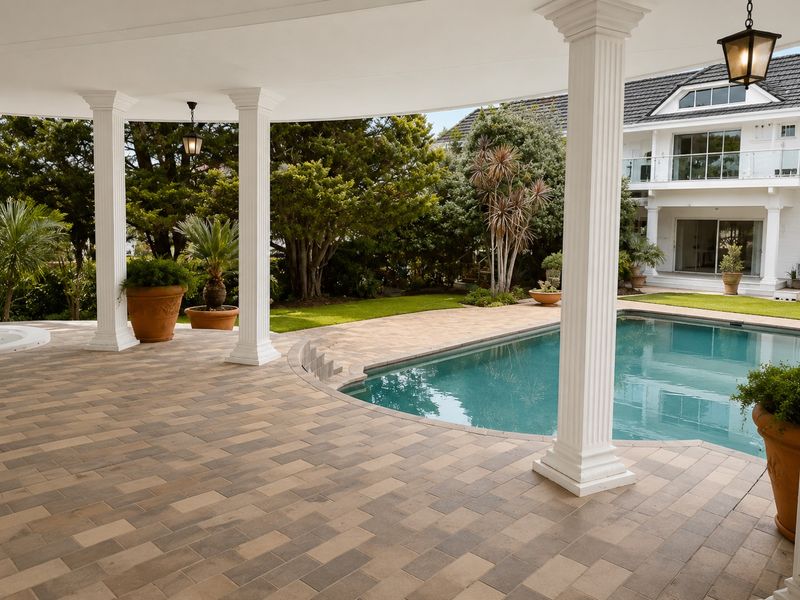 A spacious covered patio area featuring classical columns, overlooking a well-maintained swimming pool and lush garden setting.