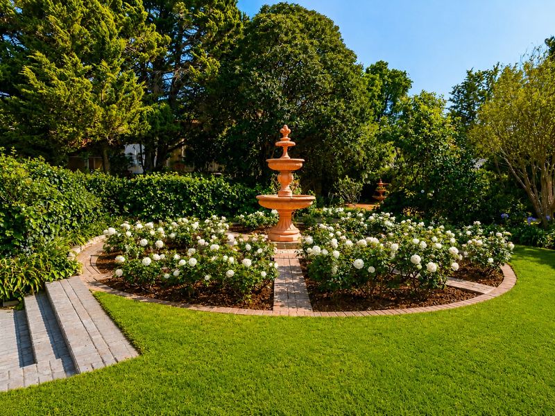 A beautifully landscaped garden featuring a central stone fountain, circular rose beds, brick pathways, and mature greenery.