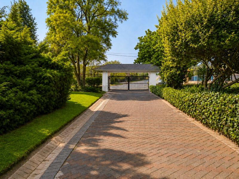 A well-maintained paved driveway leading to a secure gated entrance, framed by lush mature trees and manicured garden borders.
