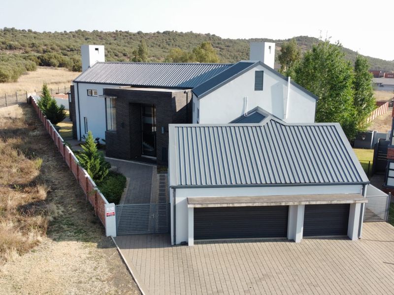 Aerial view of a modern house featuring a metal roof, double garage, paved driveway, and secure boundary walling in a scenic setting.
