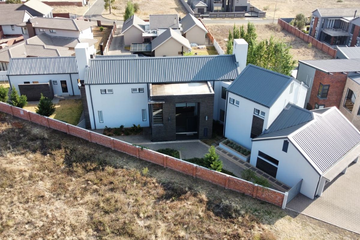 Aerial view of a modern residential property featuring a multi-pitched metal roof, brick boundary wall, and paved driveway in a suburban estate setting.