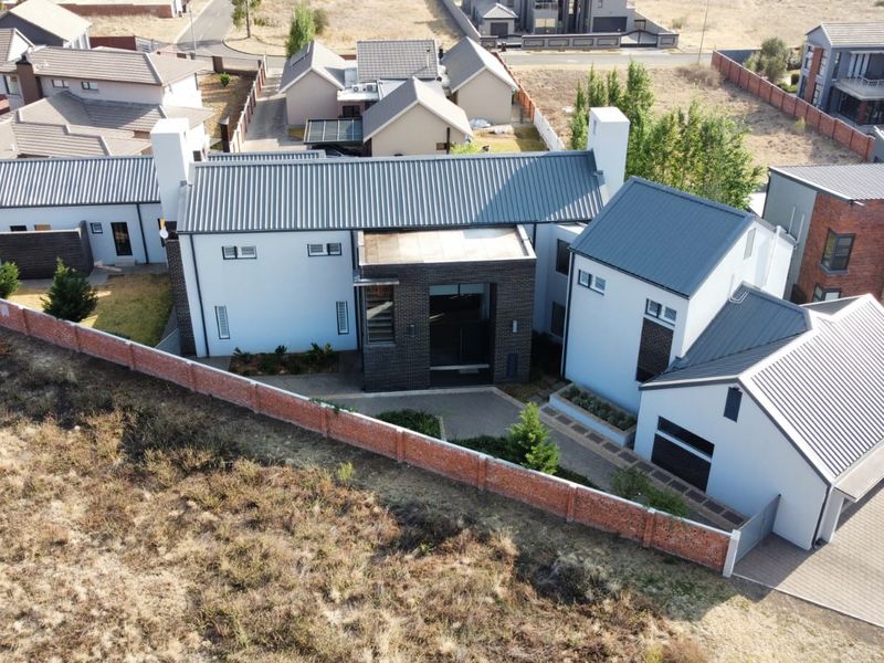 Aerial view of a modern residential property featuring a multi-pitched metal roof, brick boundary wall, and paved driveway in a suburban estate setting.