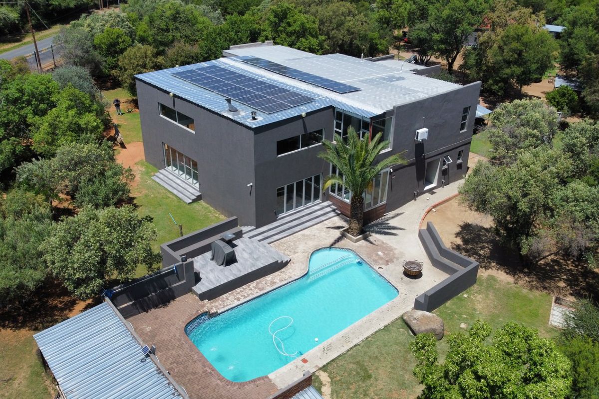 Aerial view of a modern two-story house featuring a swimming pool, paved patio area, and roof-mounted solar panels set within a landscaped garden.