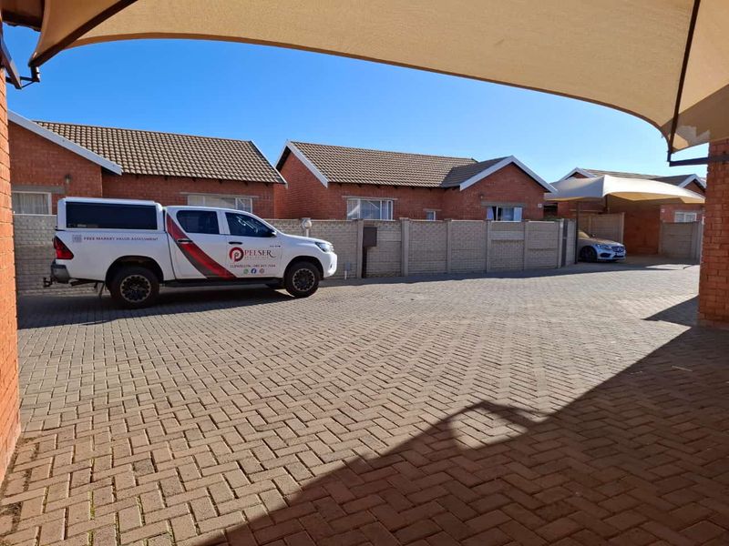 A paved driveway area featuring a shade sail covering, leading to the front facades of brick townhouse units with tiled roofs.