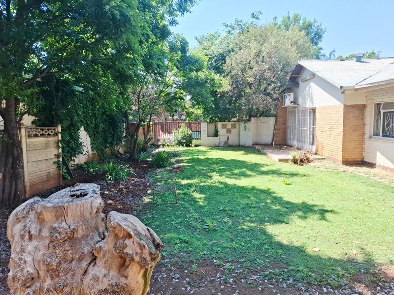 A spacious rear garden featuring a lawn area, mature trees, perimeter walling, and a paved patio section adjacent to the house.
