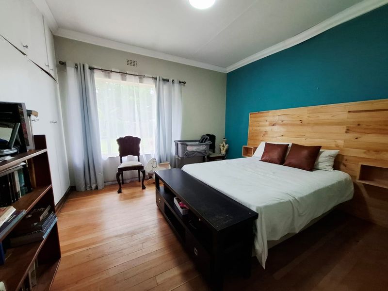 A well-proportioned bedroom featuring warm wooden flooring, built-in storage cupboards, and a decorative timber headboard against a bold accent wall.