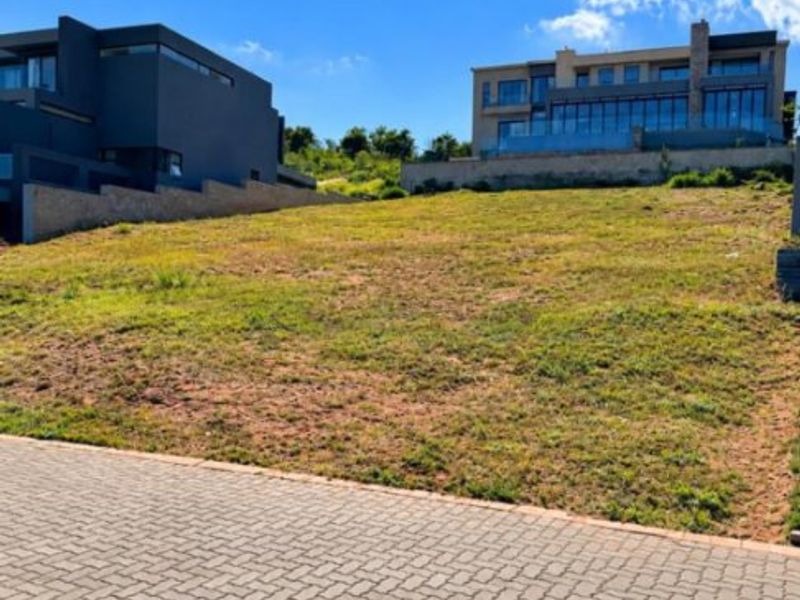 A vacant residential plot featuring a paved driveway and a gentle slope, situated in an established neighborhood with surrounding modern homes.