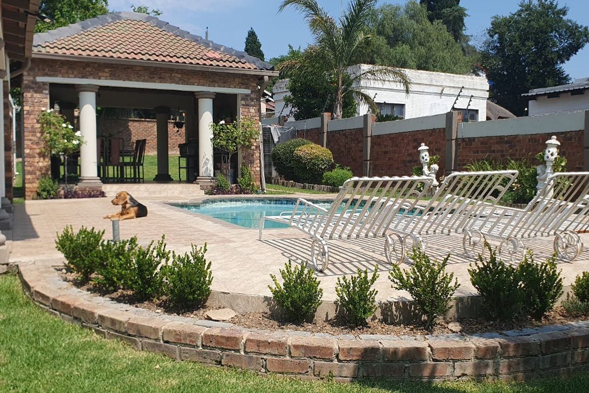 A well-maintained outdoor pool area featuring a paved deck, a covered patio with brick columns, and a landscaped garden with a brick retaining wall.