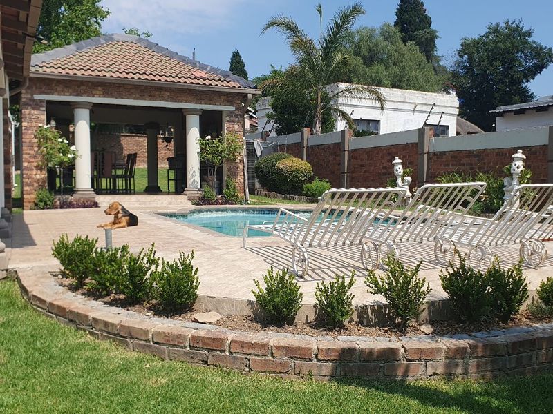 A well-maintained outdoor pool area featuring a paved deck, a covered patio with brick columns, and a landscaped garden with a brick retaining wall.