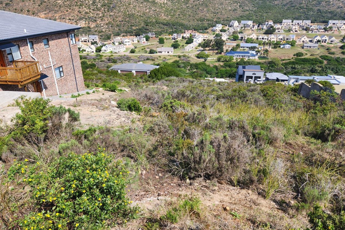 A view of a vacant residential plot featuring natural vegetation, overlooking a developed estate with surrounding hills.