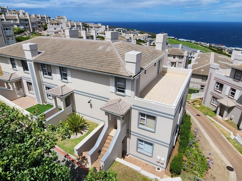 Aerial view of a residential apartment complex featuring tiled roofs, balconies, and paved walkways with a scenic coastal backdrop.