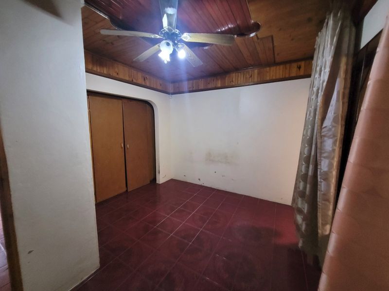 An empty bedroom featuring a timber ceiling with a ceiling fan, tiled flooring, and built-in cupboards, requiring minor wall repairs and cosmetic updates.