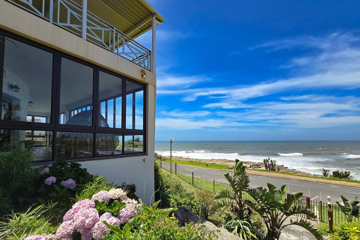A rear exterior view of the property featuring a balcony, large windows, and a landscaped garden overlooking the coastal road and ocean.