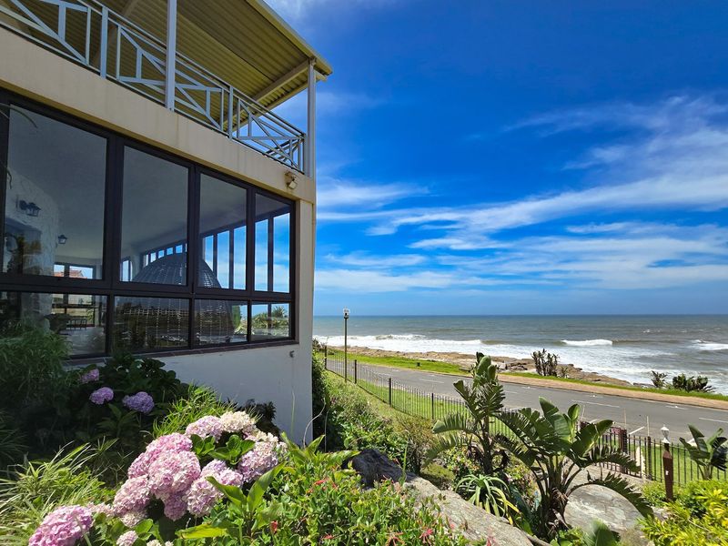 A rear exterior view of the property featuring a balcony, large windows, and a landscaped garden overlooking the coastal road and ocean.