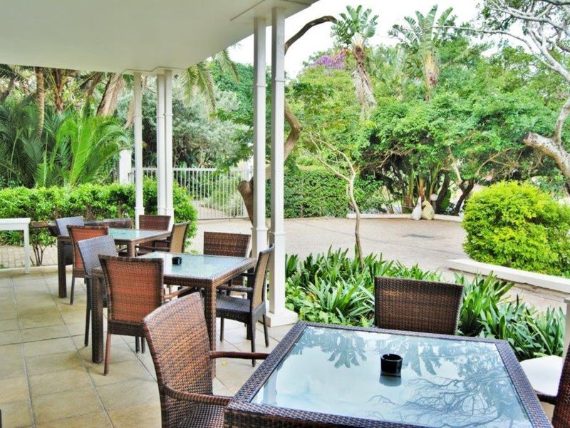 A covered outdoor patio area featuring tiled flooring and support columns, overlooking a landscaped garden and paved driveway.