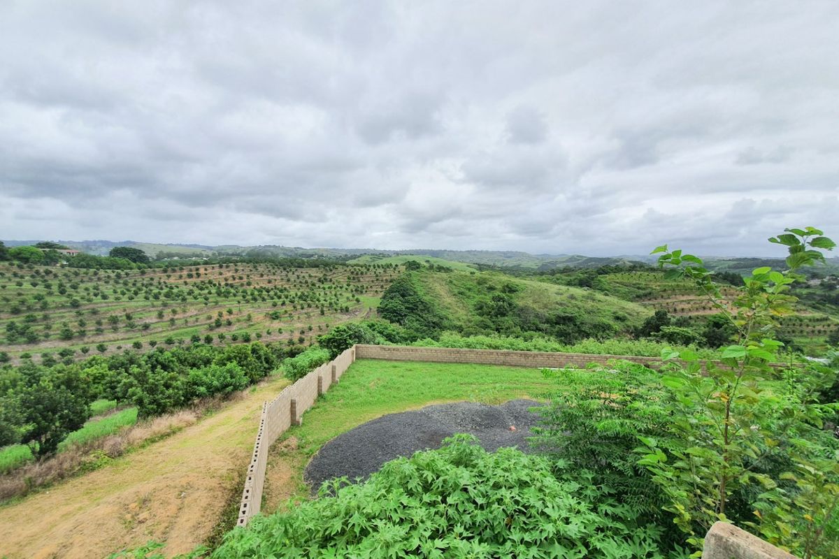 Elevated view overlooking a rural landscape with orchards, featuring a partially enclosed plot with a concrete block boundary wall and gravel area.