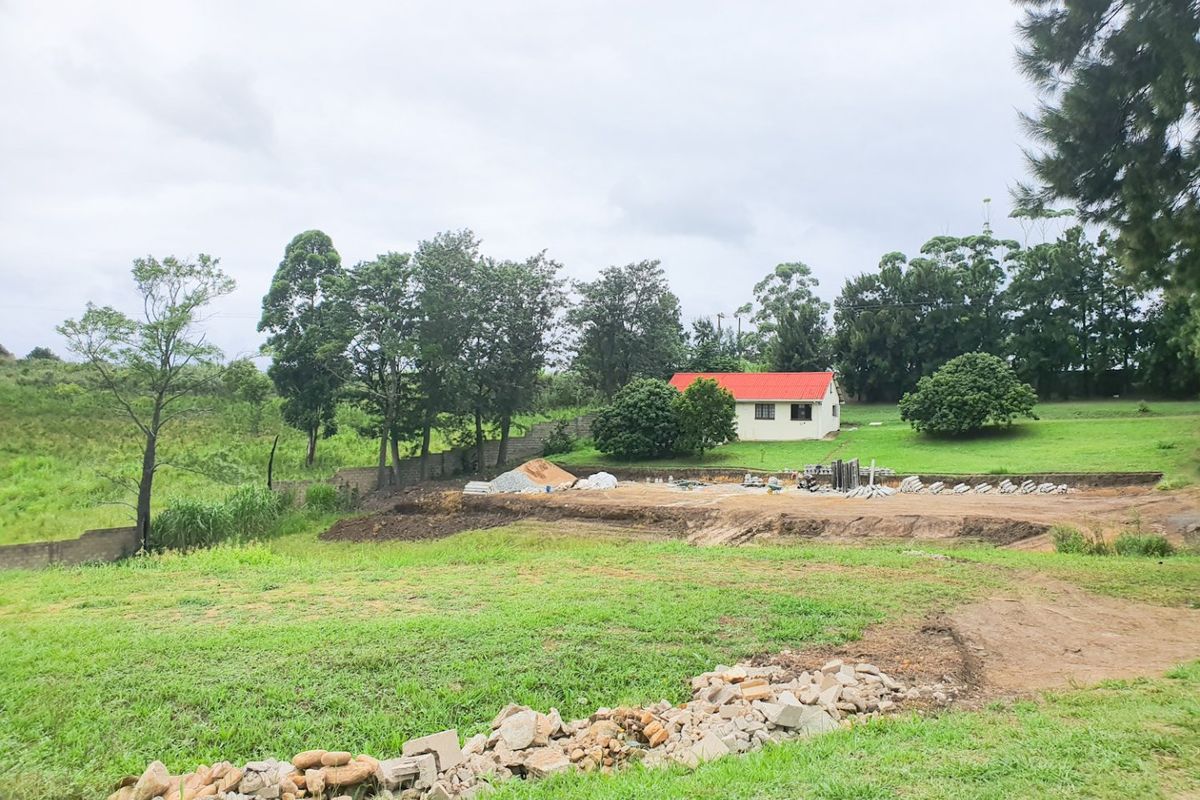 A view of the rear property grounds featuring a cleared construction area, retaining wall, and surrounding green space with a small building in the distance.