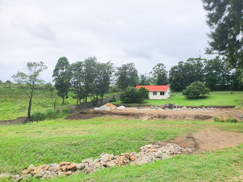 A view of the rear property grounds featuring a cleared construction area, retaining wall, and surrounding green space with a small building in the distance.
