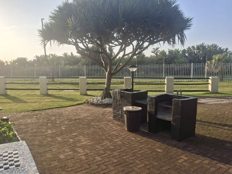 Outdoor patio area featuring a built-in brick braai, paved flooring, and established garden surroundings within a fenced complex.