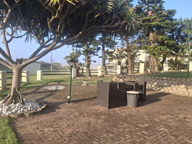 A communal garden area featuring brick paving, a built-in braai, and established trees within a fenced apartment complex.