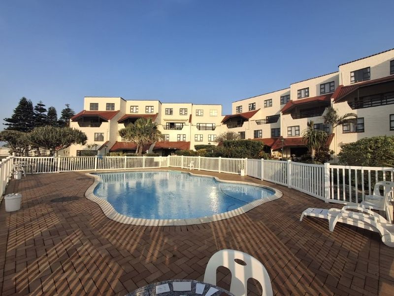 A well-maintained communal swimming pool area featuring paved decking and secure perimeter fencing, set against a multi-story apartment complex.