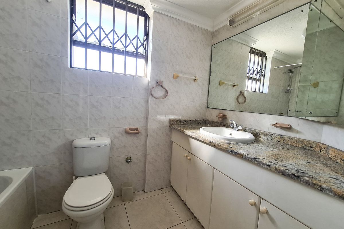 A functional bathroom featuring a granite-topped vanity, wall-mounted mirror, toilet, and tiled walls with a window protected by security bars.