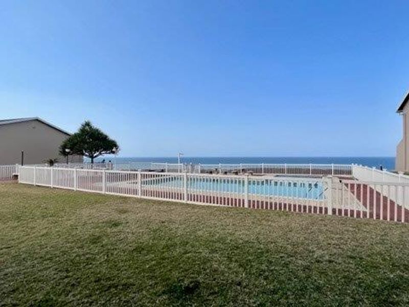 Communal swimming pool area featuring a paved surround, safety fencing, and expansive lawn with ocean views.
