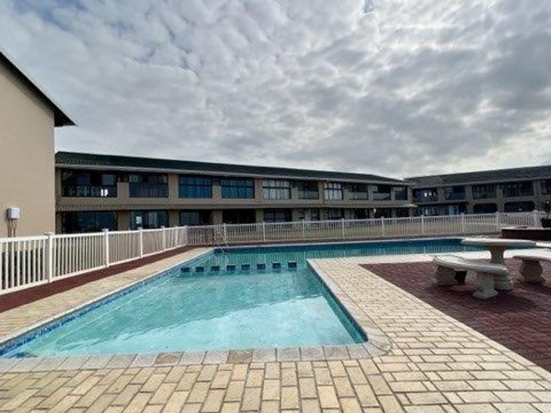 A well-maintained communal swimming pool area featuring paved surrounds and a white picket fence, set within an apartment complex exterior.