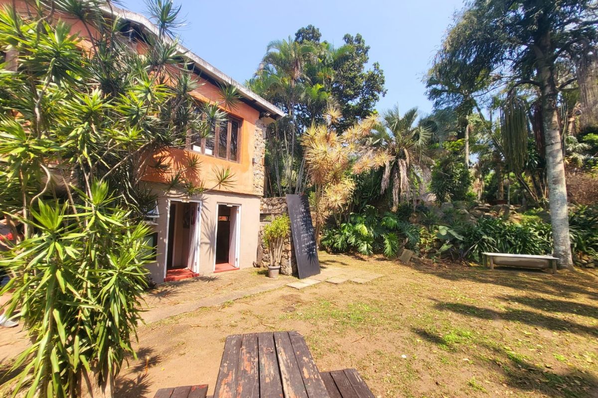 A rear view of a rustic farm-style building featuring stone and plastered walls, set within a lush, established garden area.