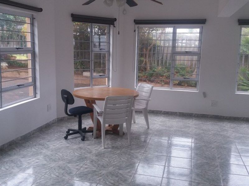 A bright, open living area featuring tiled flooring, large windows with security bars, and a ceiling fan, offering a clean and functional space.