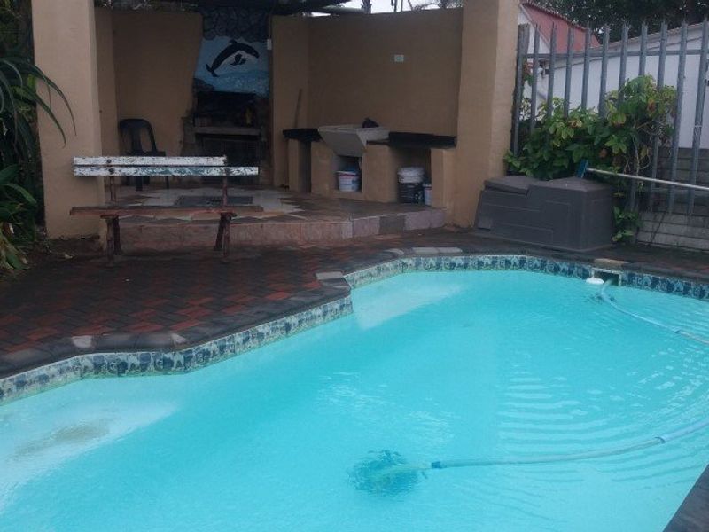 Outdoor pool area featuring a covered patio with a built-in braai, concrete sink, and brick paved surround, requiring some maintenance and cosmetic updates.