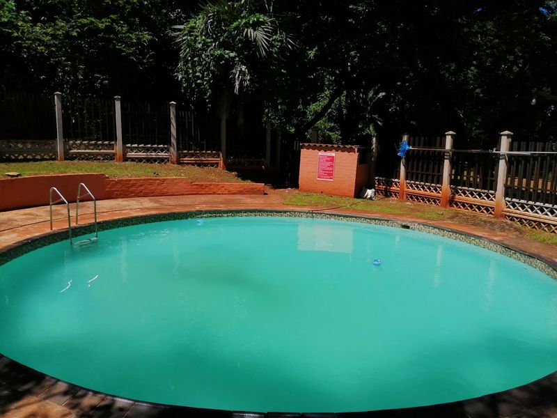 A circular swimming pool area featuring a brick retaining wall and perimeter fencing, set within a shaded garden environment.
