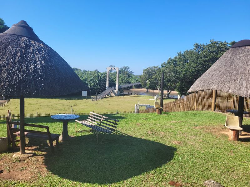 Scenic outdoor area featuring thatched gazebos overlooking a lush green lawn, river, and pedestrian suspension bridge.
