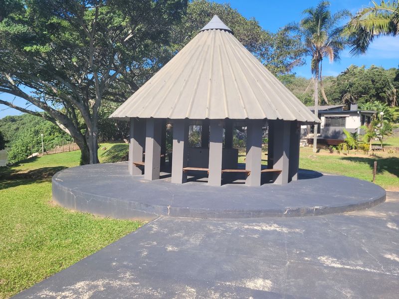 A circular outdoor gazebo or lapa featuring a conical roof, concrete pillars, and built-in bench seating, set within a landscaped garden area.
