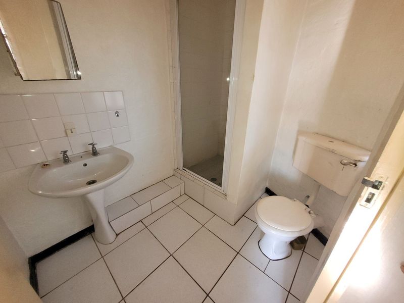 A dated bathroom featuring a pedestal basin, shower enclosure, and toilet with white tiled flooring and splashbacks, requiring cosmetic updates.
